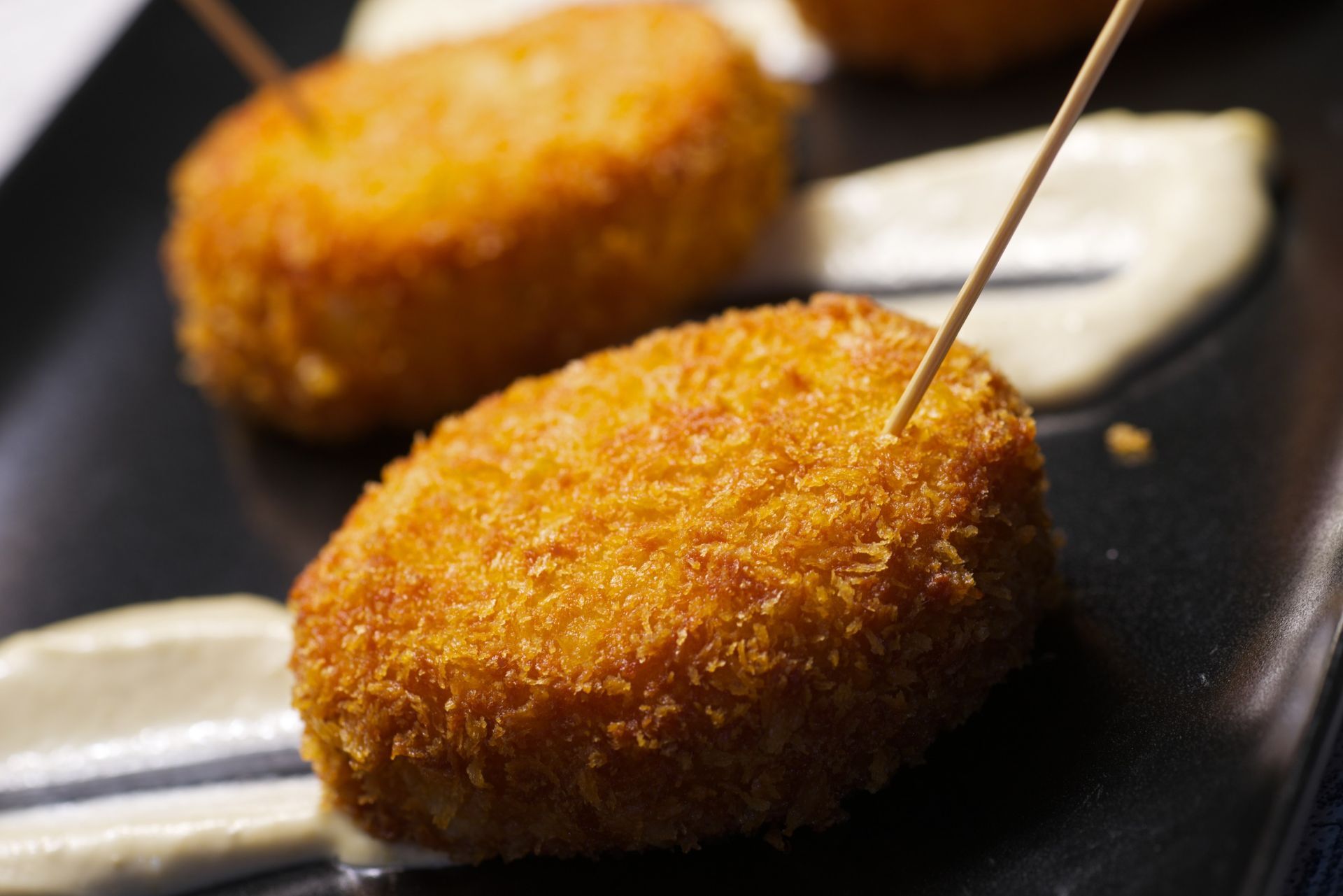 Primer plano de croquetas doradas y empanizadas en un plato oscuro, cada una con un palillo y un remolino de salsa blanca.