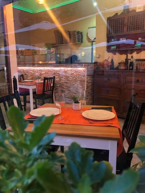 Interior de una pizzería, visto a través de una ventana. Una mesa está puesta con platos y vasos, y al fondo se ve una zona de preparación de pizzas.