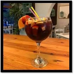 Una copa grande de vino llena de sangría roja, adornada con frutas y pajitas, reposa sobre una mesa de madera.