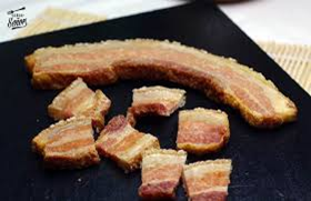 Primer plano de panceta de cerdo crujiente, entera y cortada en cubos, sobre una tabla de cortar negra. La carne está dorada y tiene capas de carne y grasa.