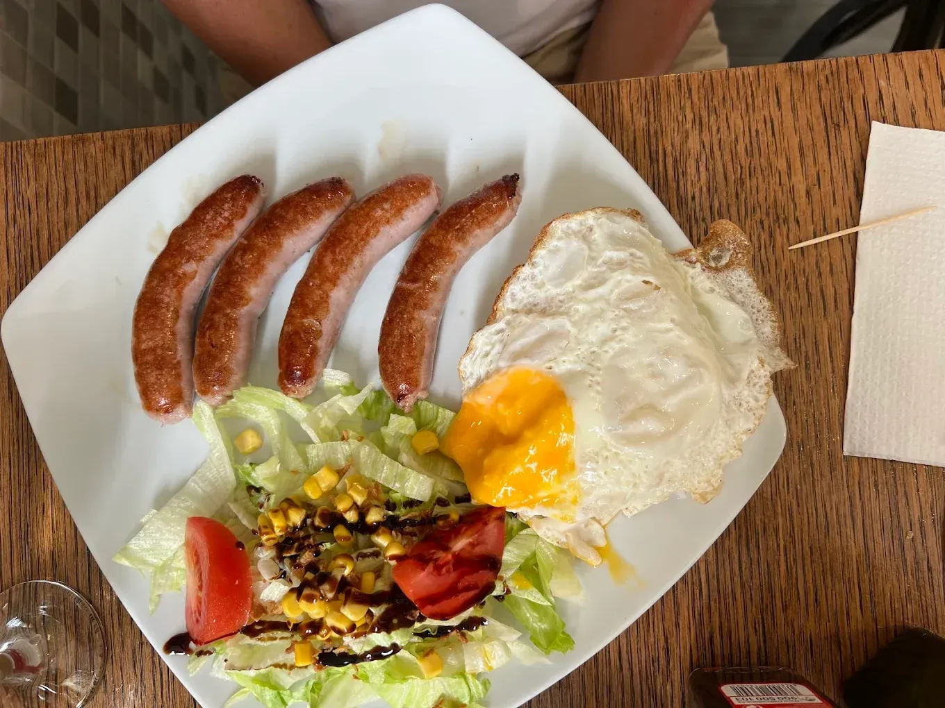 Plato con cuatro salchichas, huevo frito y una pequeña ensalada con maíz y tomate sobre una mesa de madera.