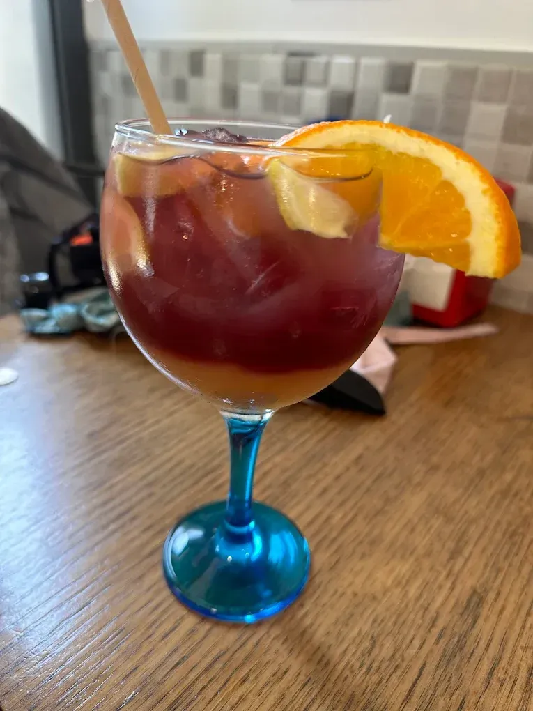 Un colorido cóctel de sangría en una copa de tallo azul adornada con una rodaja de naranja y una pajita, sobre una mesa de madera.