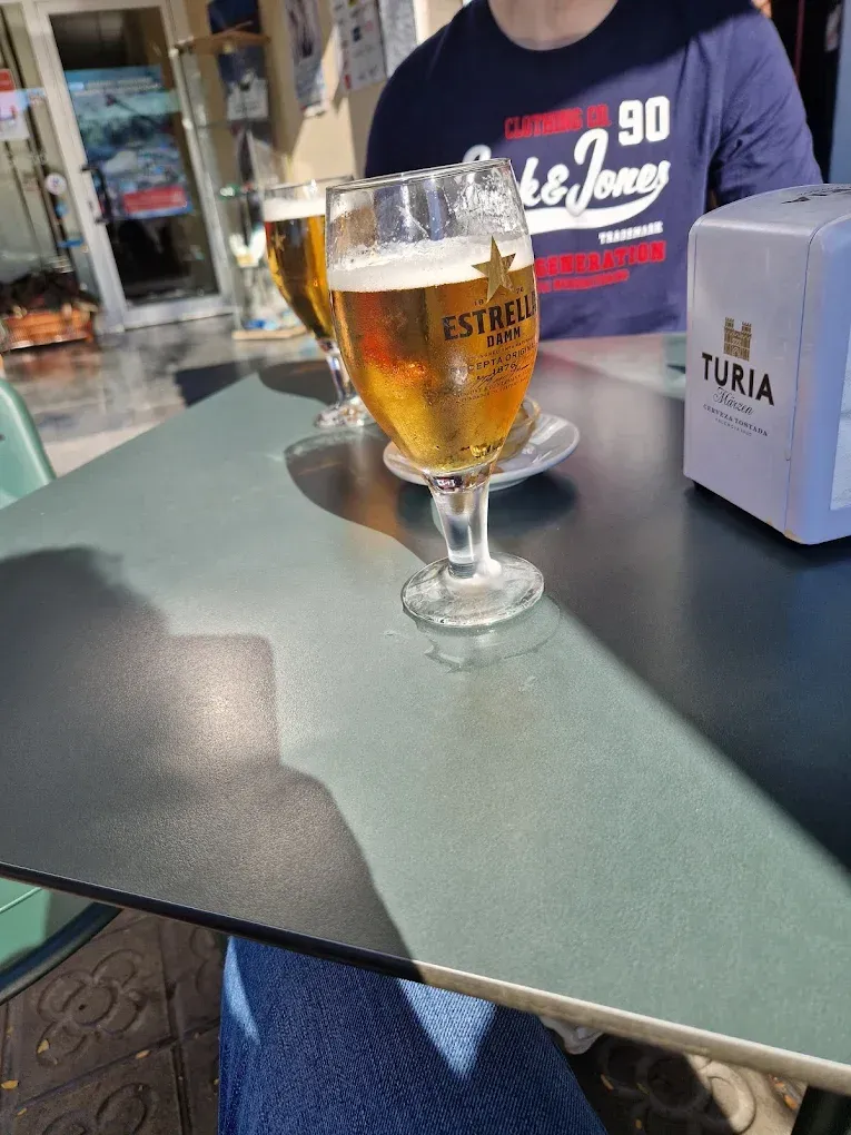 Dos vasos de cerveza Estrella sobre una mesa, con una persona con camisa azul al fondo. Un servilletero cerca.