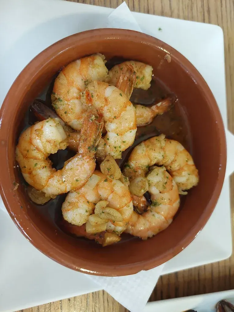Un pequeño cuenco de barro lleno de camarones cocidos nadando en un aceite sabroso, adornado con hierbas y ajo.