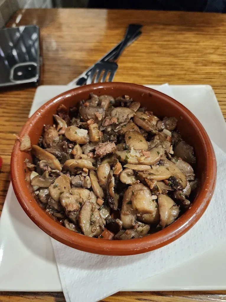 Hongos en un recipiente de cerámica marrón sobre un plato cuadrado blanco, con tenedores en el fondo.