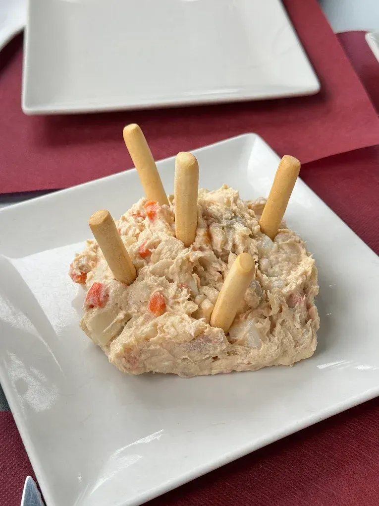 Un montículo de comida beige y cremosa en un plato cuadrado blanco, adornado con palitos de pan. Servido sobre un mantel burdeos.