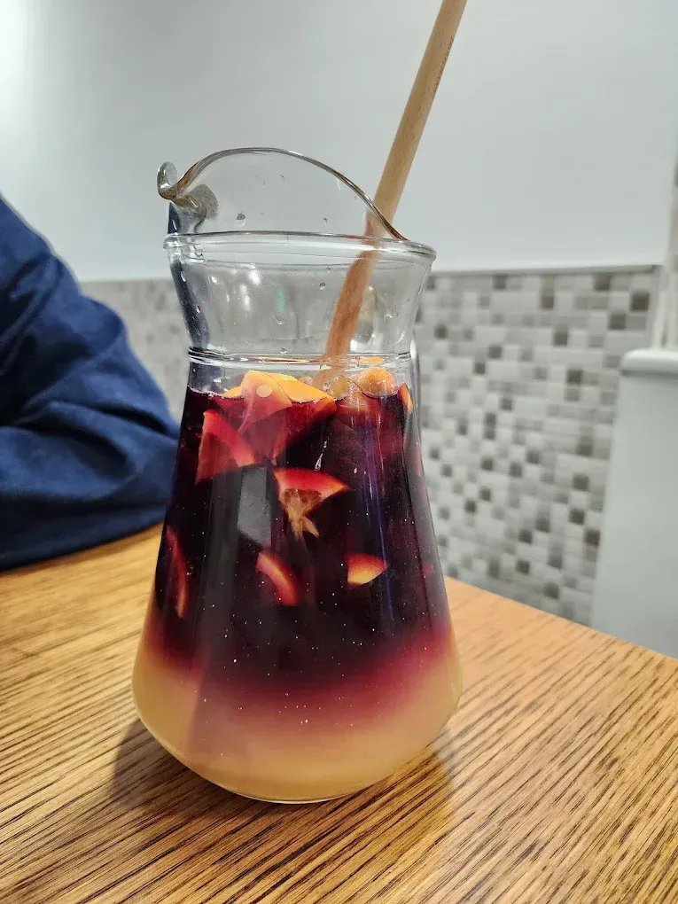 Una jarra de sangría con rodajas de fruta y una pajita de madera sobre una mesa de madera. La bebida presenta un degradado de rojo y amarillo.