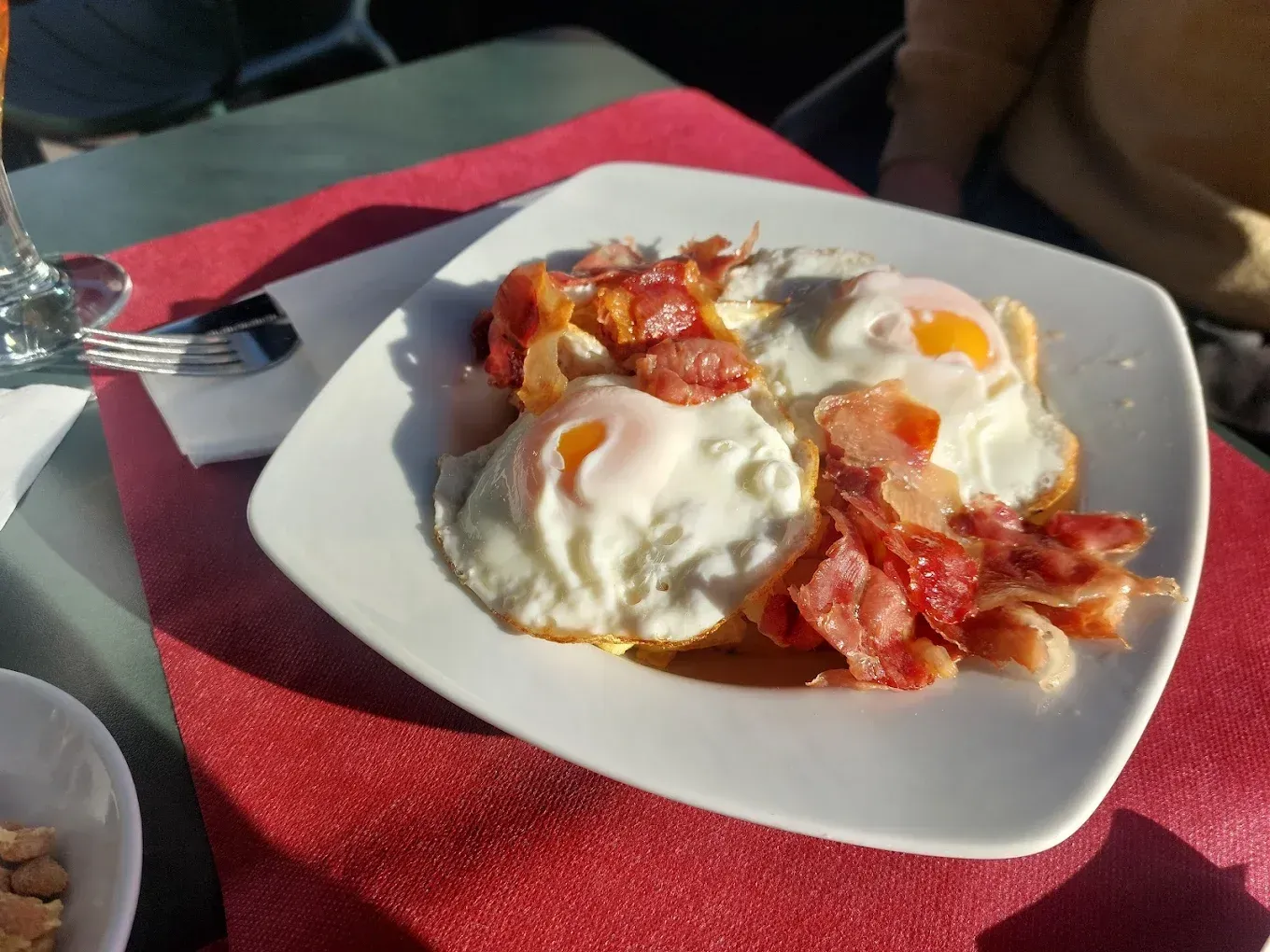 Huevos fritos con tocino y tomate en un plato blanco, sobre un mantel rojo. Luz solar brillante.