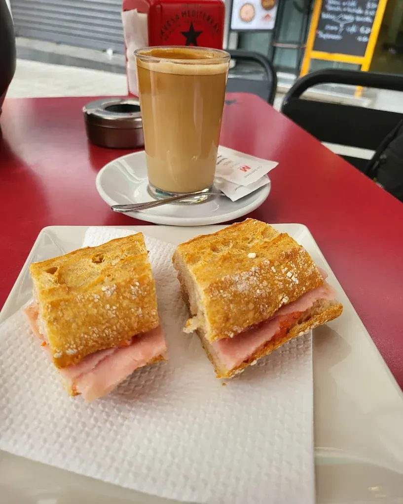 Sándwich y café sobre una mesa roja. Se ven una baguette cortada por la mitad, un vaso de café y un cenicero.