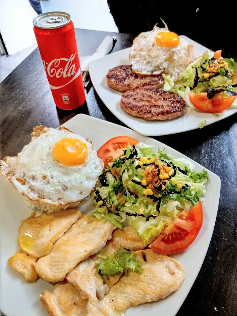 Dos platos de comida con huevos fritos, empanadas de carne y ensalada, junto con una lata de Coca-Cola sobre una mesa oscura.