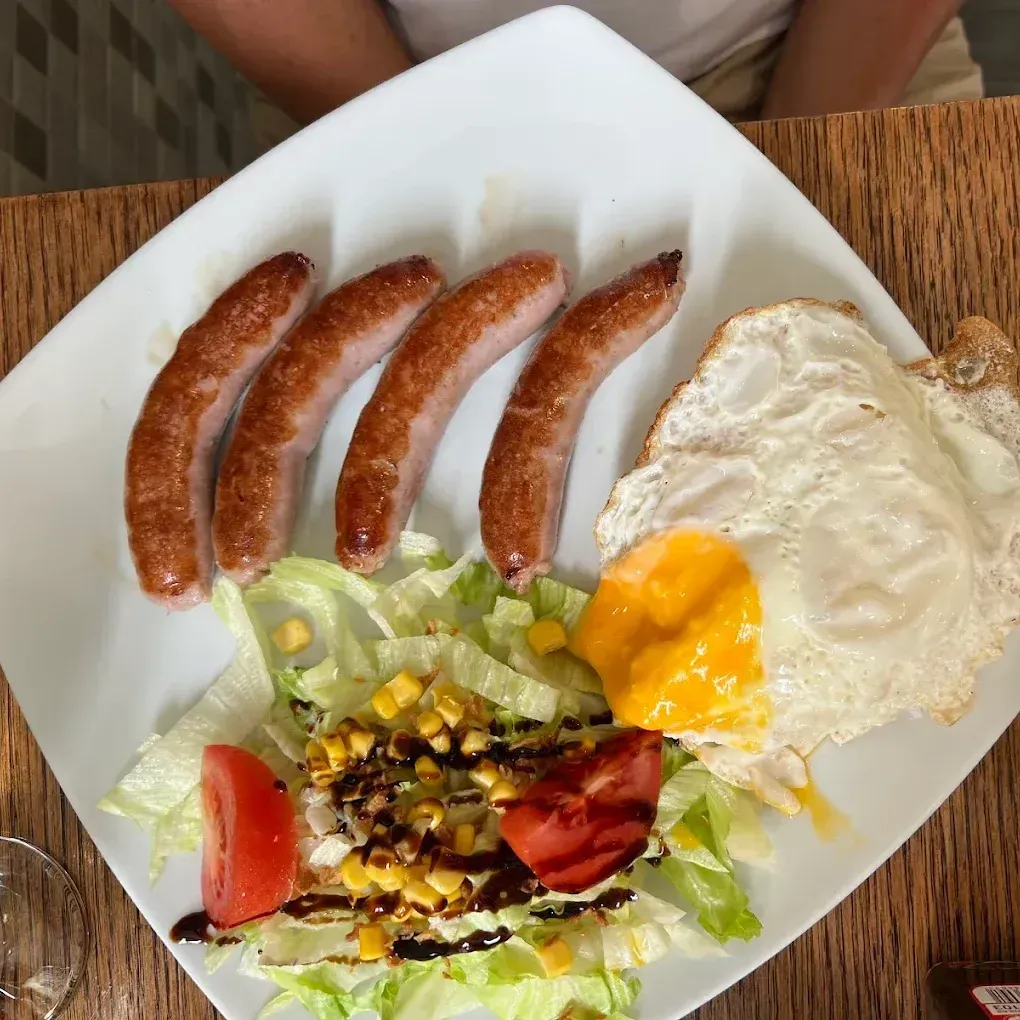 Plato de comida: cuatro salchichas, huevo frito, ensalada con maíz y tomate.