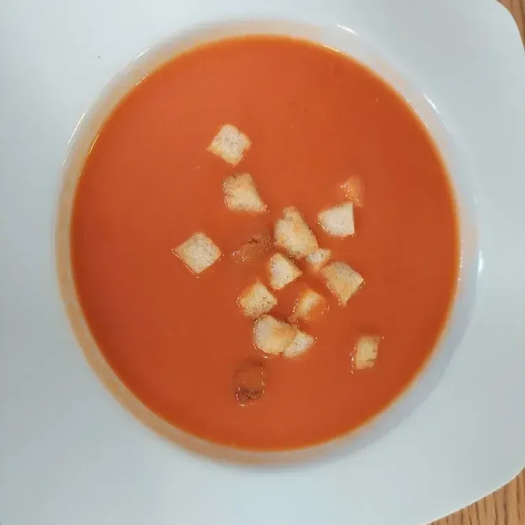 Sopa de tomate en un tazón blanco, con crutones encima. La sopa es de un vibrante color naranja rojizo.