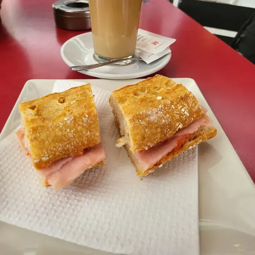 Dos mitades de un sándwich de jamón en un plato blanco, con una bebida de café de fondo sobre una mesa roja.