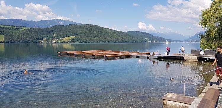 Ein Mann steht auf einem Steg neben einem See mit Bergen im Hintergrund.