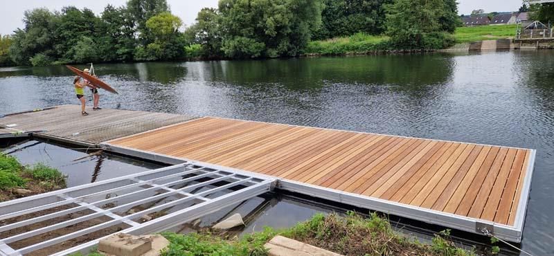 Eine Person steht auf einem Holzsteg und hält ein Surfbrett.