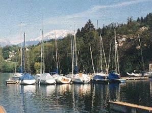 Eine Reihe Segelboote liegen in einem Hafen vor Anker, im Hintergrund sind Berge zu sehen.