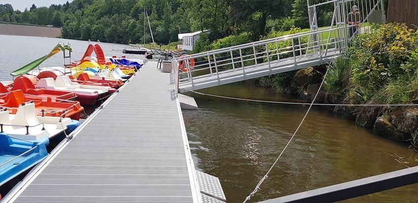 Eine Reihe Tretboote liegen an einem Steg neben einem Fluss.