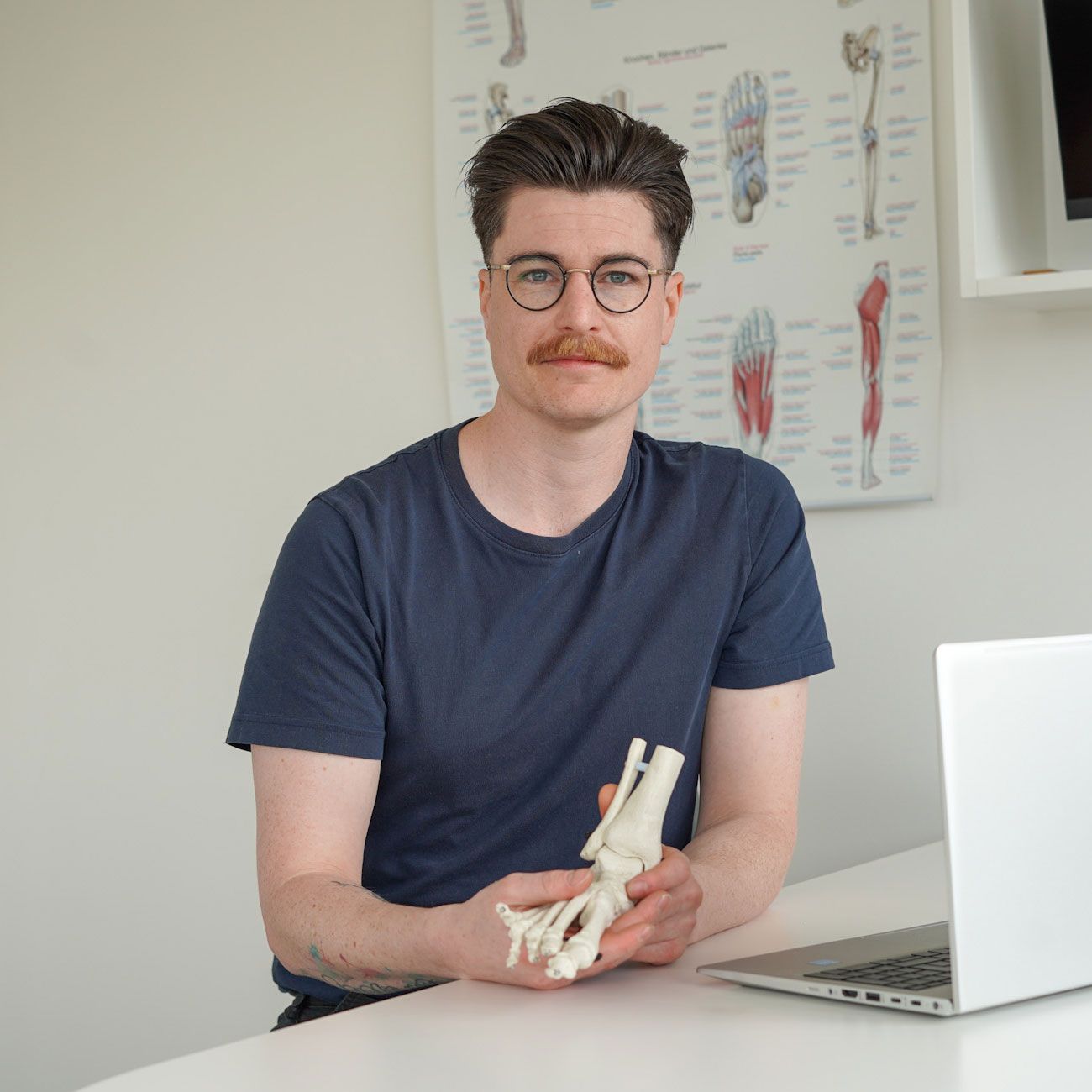 Eine Person mit Brille und Schnurrbart sitzt an einem Schreibtisch und hält ein anatomisches Modell eines menschlichen Fußes in der Hand; neben ihr steht ein Laptop.
