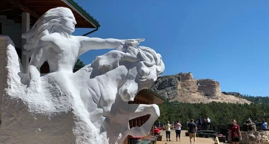 Large white snow sculpture with people walking below, set against a sunny sky and rocky backdrop