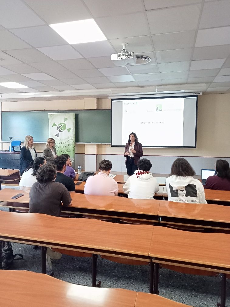 Un presentador se sitúa frente a un grupo de estudiantes en un aula, señalando una pantalla de proyección, mientras otras dos personas permanecen de pie cerca.