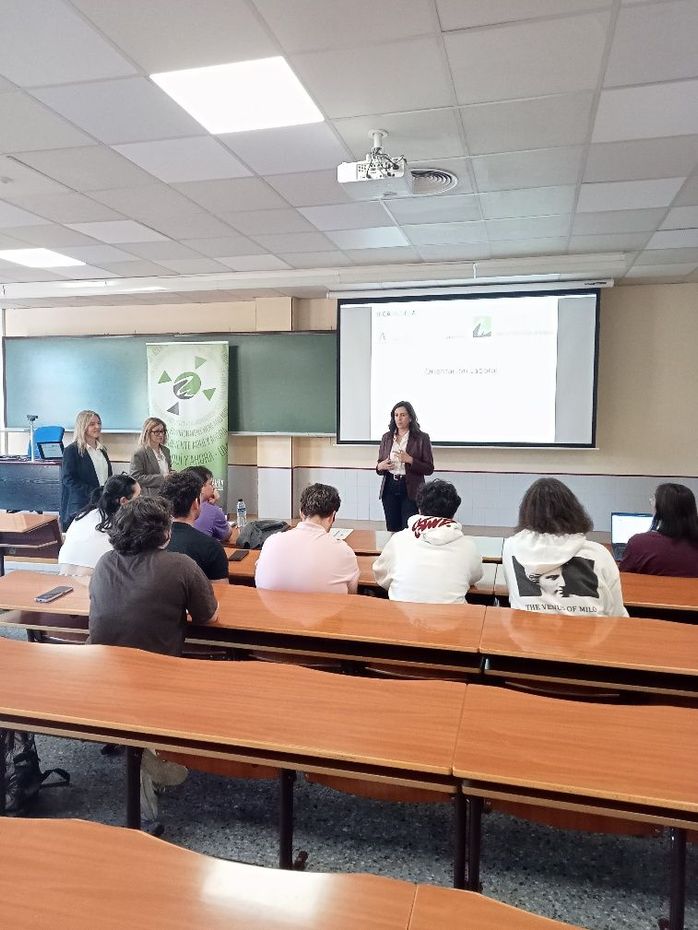 Clase magistral con los alumnos sentados en sus pupitres y un presentador hablando junto a una pantalla proyectada.