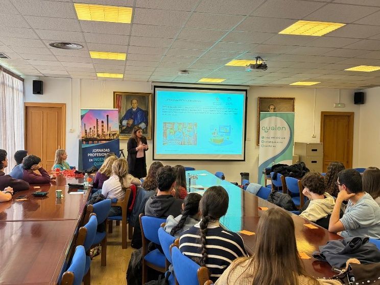 Un presentador se sitúa frente a una pantalla de proyección en un aula, dirigiéndose a un público sentado en largas mesas.
