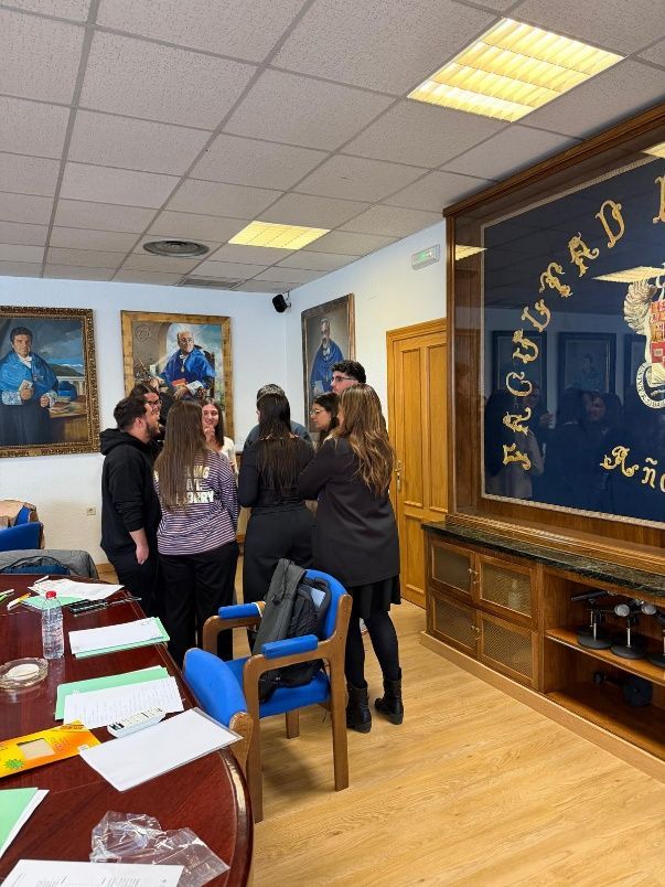 Un pequeño grupo se encuentra en una sala con retratos y una gran vitrina con marcos negros y dorados, reunidos cerca de sillas azules.