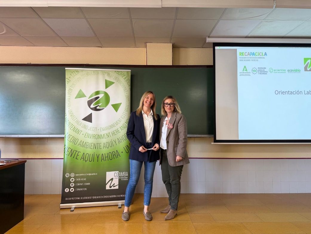 Dos personas están de pie una al lado de la otra frente a una pancarta verde del evento y una pantalla de proyección en un aula.