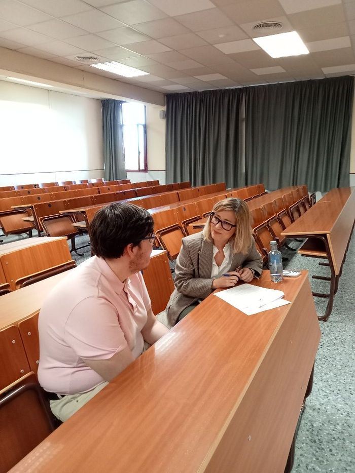 Dos personas conversando en un escritorio en un aula de conferencias, con filas de asientos de madera y cortinas cerradas al fondo.
