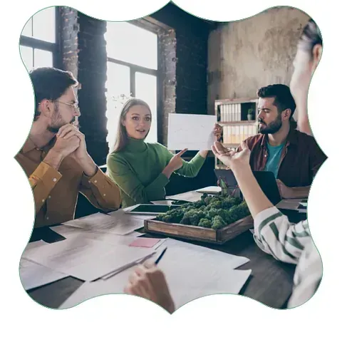 Cuatro personas en una mesa colaboran en un proyecto. La mujer señala una maqueta. La habitación es una oficina soleada.