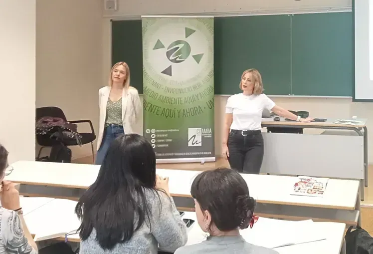 Dos mujeres dan una presentación en un aula, de cara al público. Una gesticula, con una pancarta al fondo.