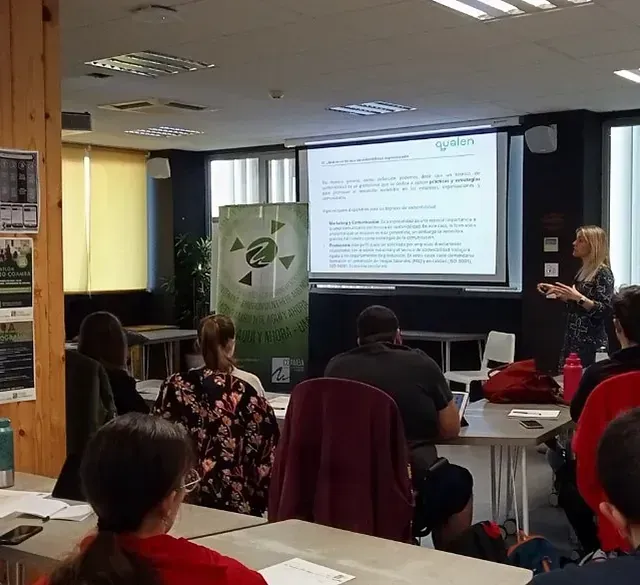 Mujer presentando en un seminario con una pantalla de proyección; audiencia sentada en mesas.