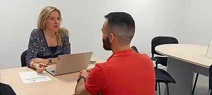 Mujer y hombre en una reunión en una mesa, mujer hablando, hombre mirándola, computadora portátil, entorno de oficina.