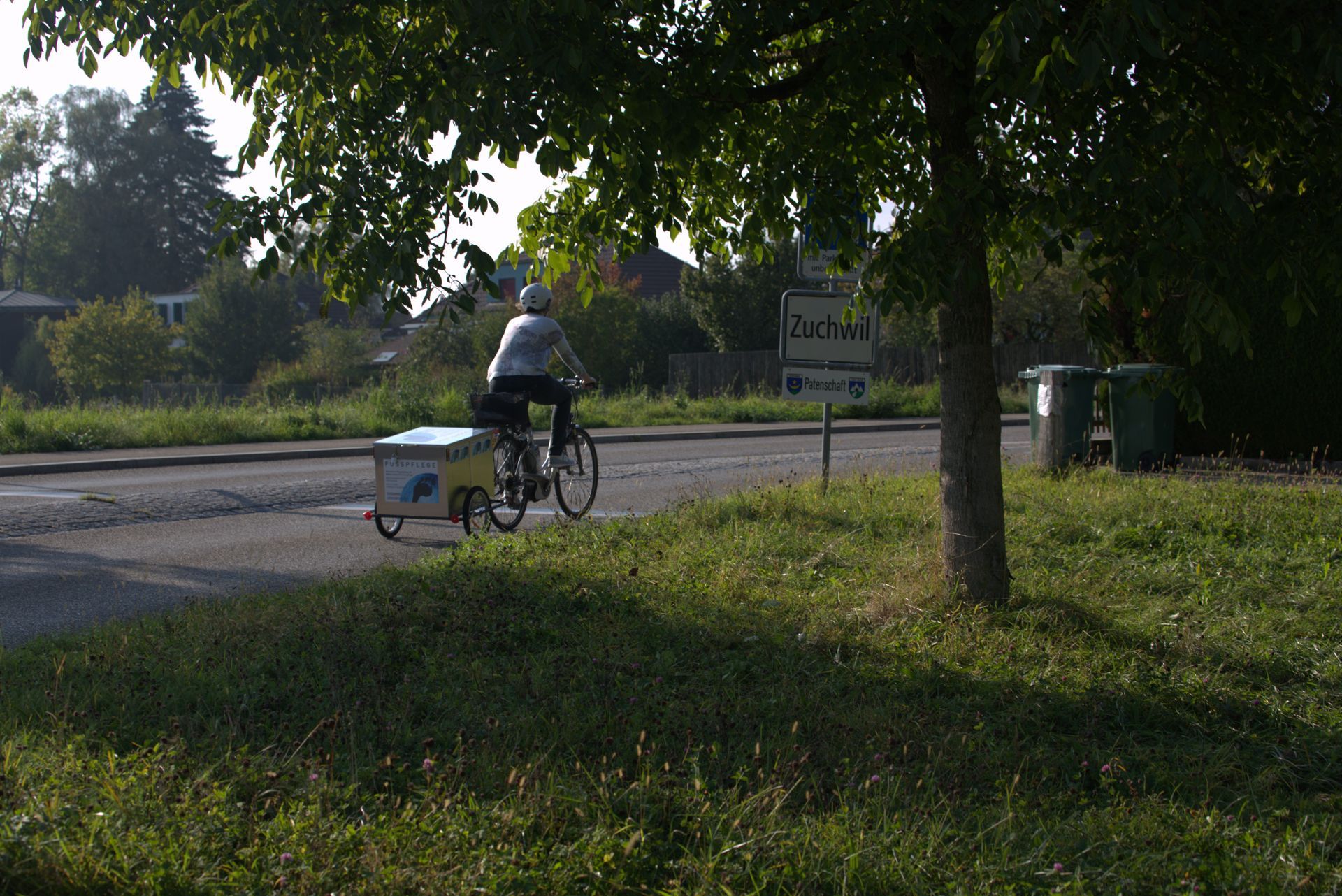 Eine Person fährt mit einem Fahrradanhänger auf einer Landstrasse neben Bäumen und Gras.