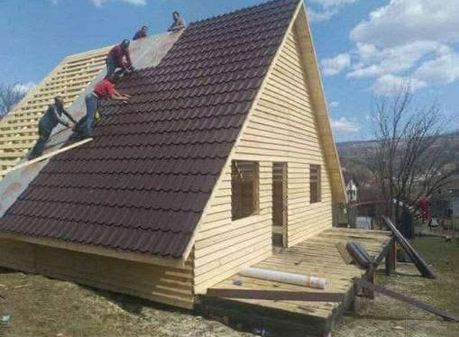 Los trabajadores de la construcción instalan un techo de metal de color marrón oscuro en una cabaña con estructura en forma de A y revestimiento de madera clara.