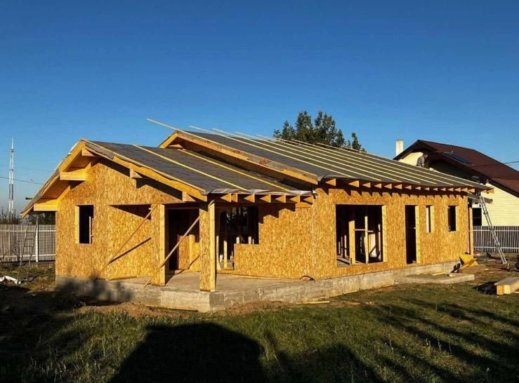 Casa en construcción: estructura de madera, paredes expuestas, techo parcialmente revestido de tejas, entorno exterior con cielo azul.