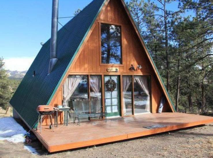 Cabaña con estructura en forma de A, revestimiento de madera marrón, techo verde y gran terraza frontal en un entorno boscoso.