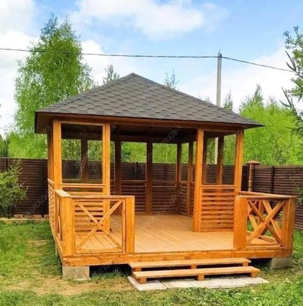 Cenador de madera con techo de tejas, sobre césped. Incluye barandilla, escalones y laterales abiertos.