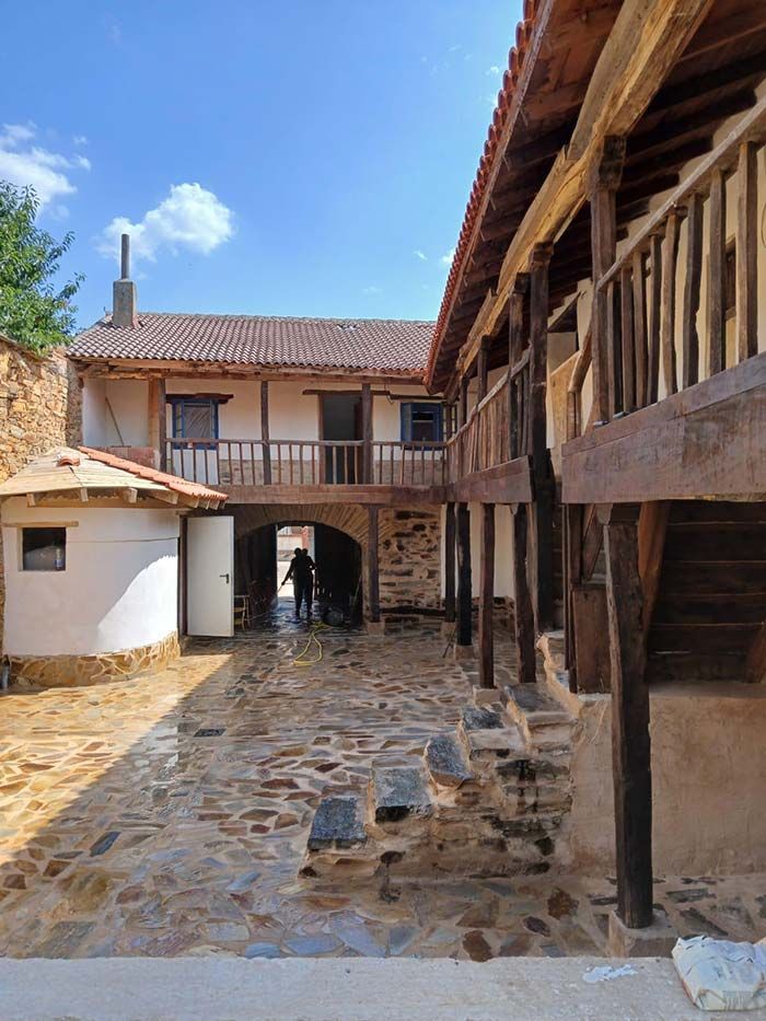 Patio con camino de piedra, balcones de madera y arco que conduce a un edificio bajo un cielo azul.