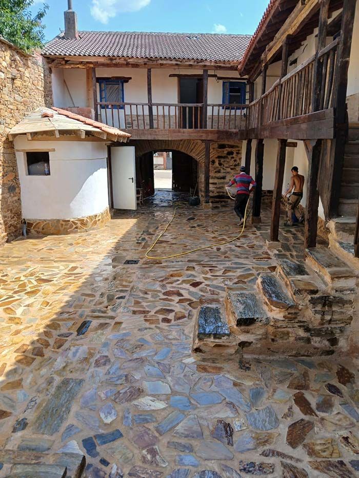 Patio con suelo empedrado, construcciones de piedra con balcones de madera y dos personas limpiando.