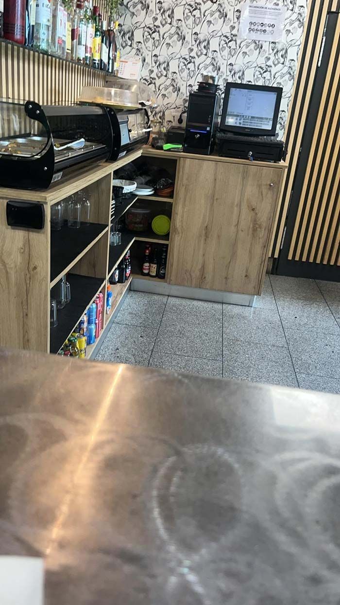 Zona de barra de bar con sistema POS, estanterías y botellas, cerca de una pared estampada.