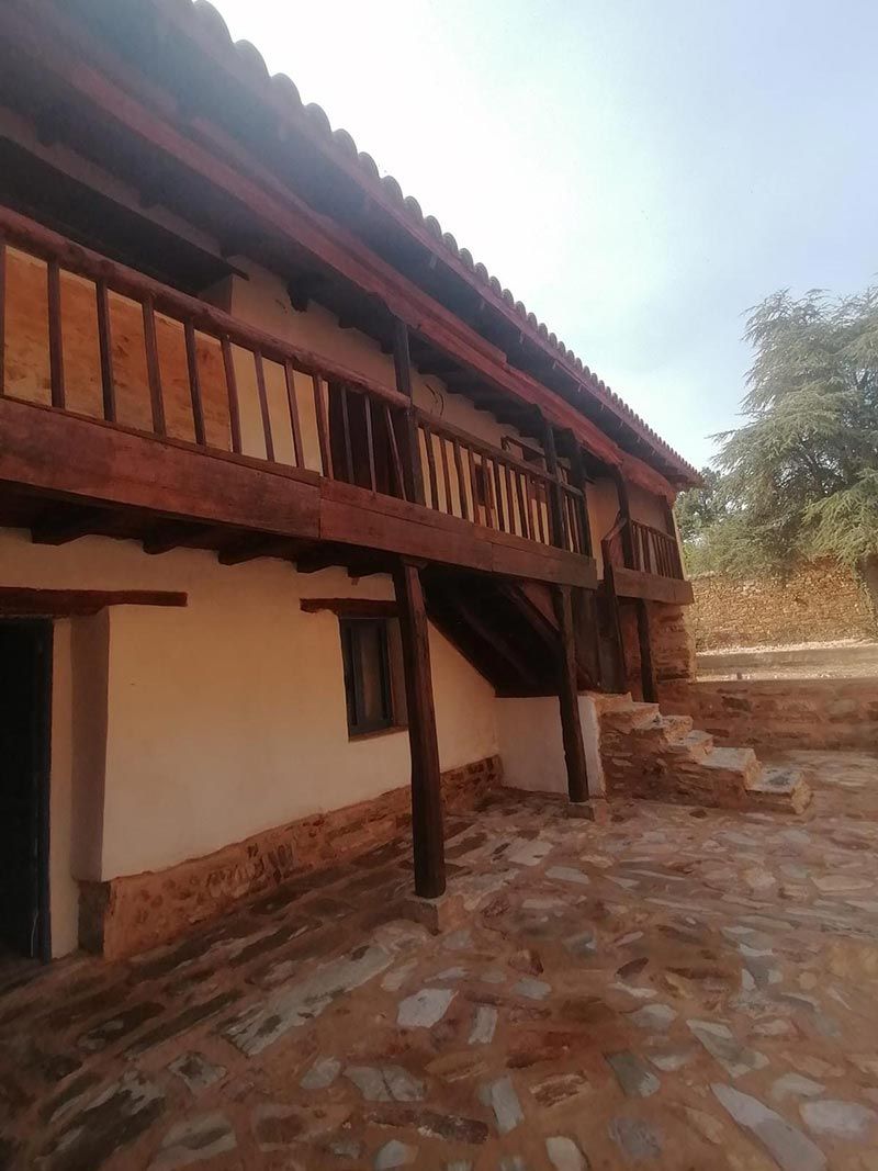 Edificio rústico de dos plantas con balcón y pilares de madera, paredes de yeso blanco y camino de piedra.