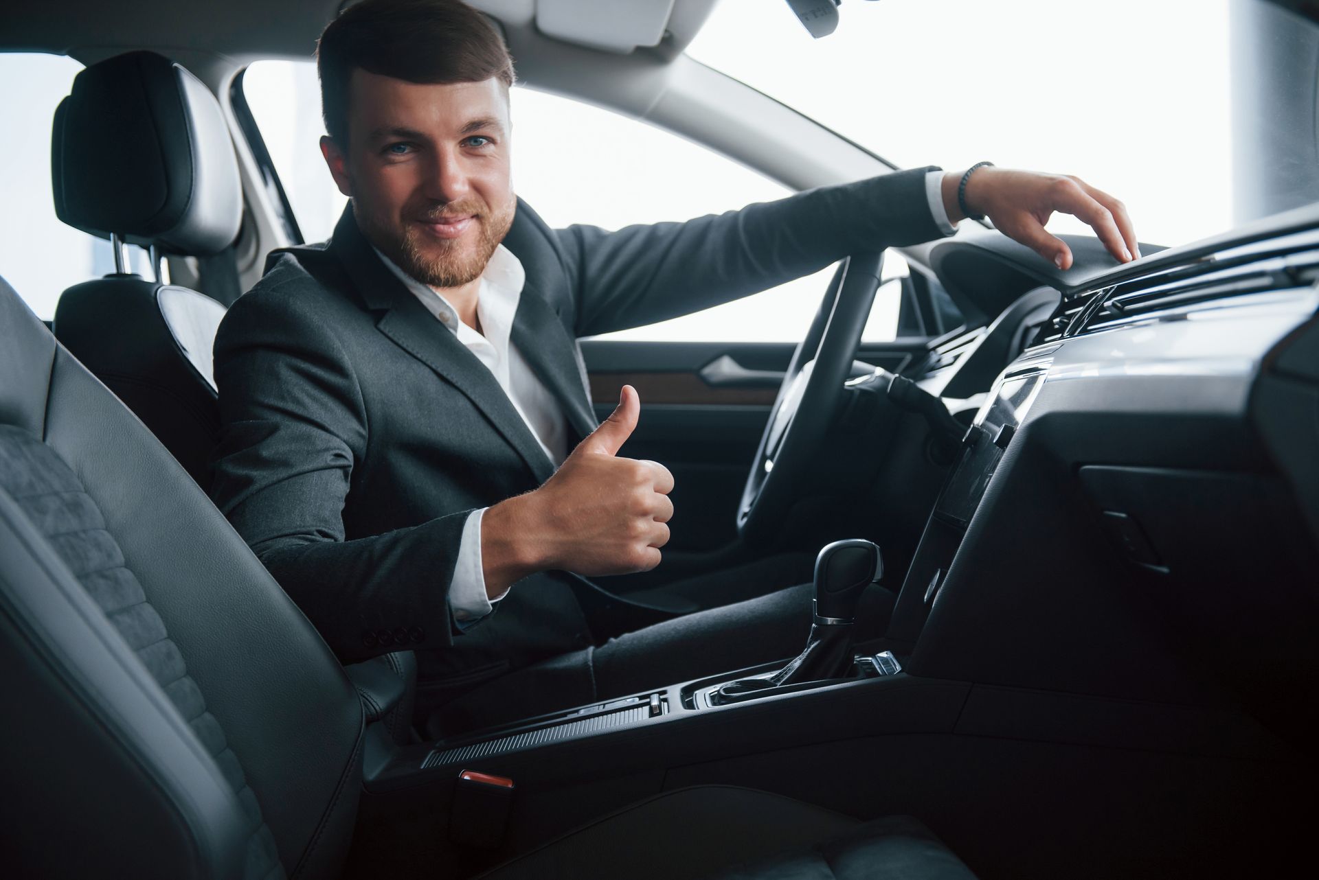 Un homme souriant en costume fait un signe de pouce levé depuis l'intérieur d'une voiture