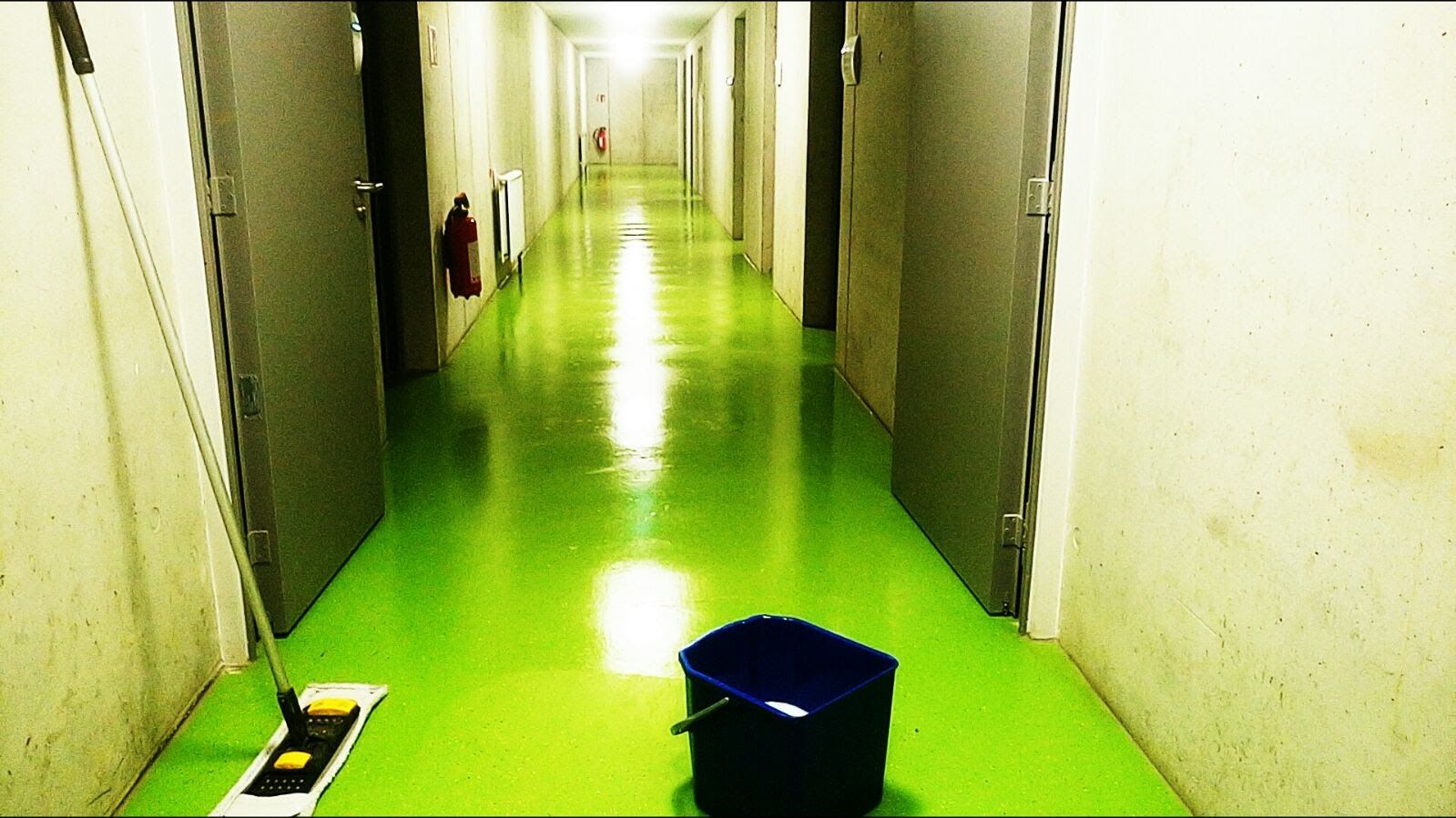 Ein langer, grüner Flur mit geschlossenen Türen; im Vordergrund Reinigungsmittel.