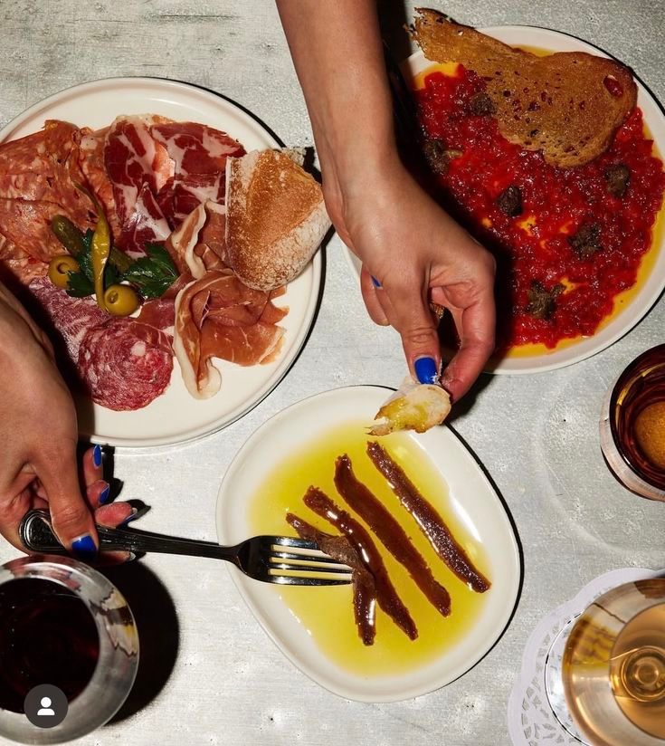 Platos de embutidos, plato rojo y anchoas con pan, manos que se extienden, escena de comedor.