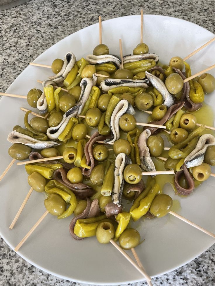 Aperitivos en brochetas: aceitunas, pimientos encurtidos y anchoas en un plato blanco.