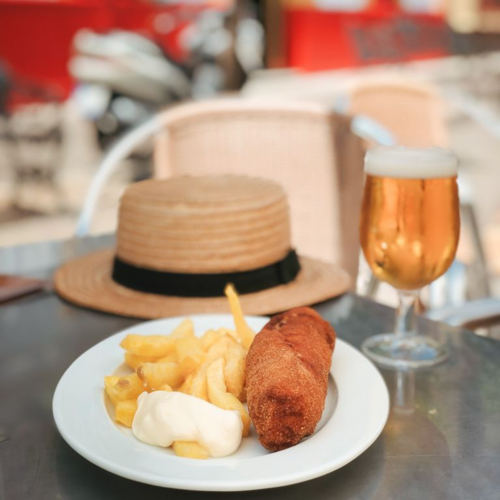 Un plato de patatas fritas y una croqueta, un sombrero y un vaso de cerveza sobre una mesa en el exterior.