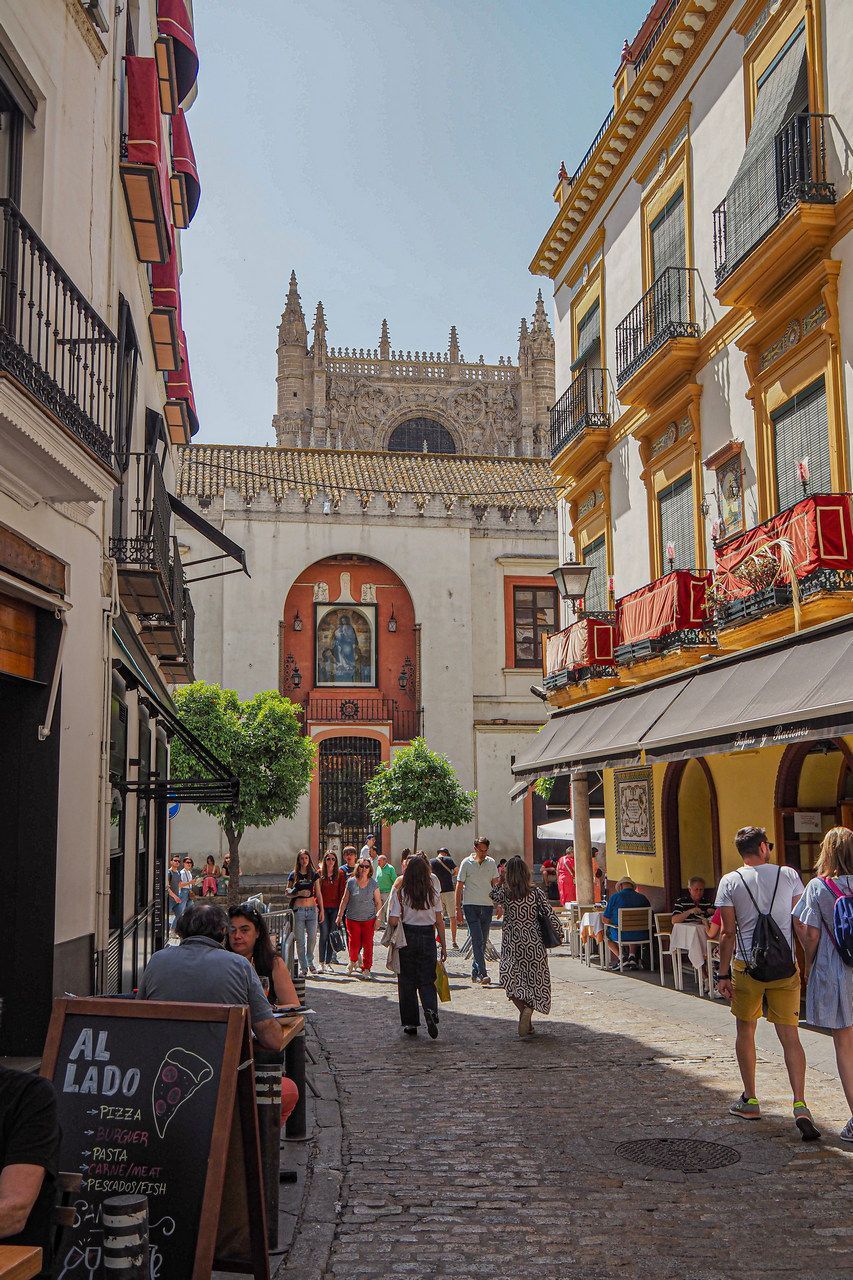 Escena callejera empedrada en Sevilla, España, con gente caminando hacia una iglesia. Terrazas de cafés y edificios
