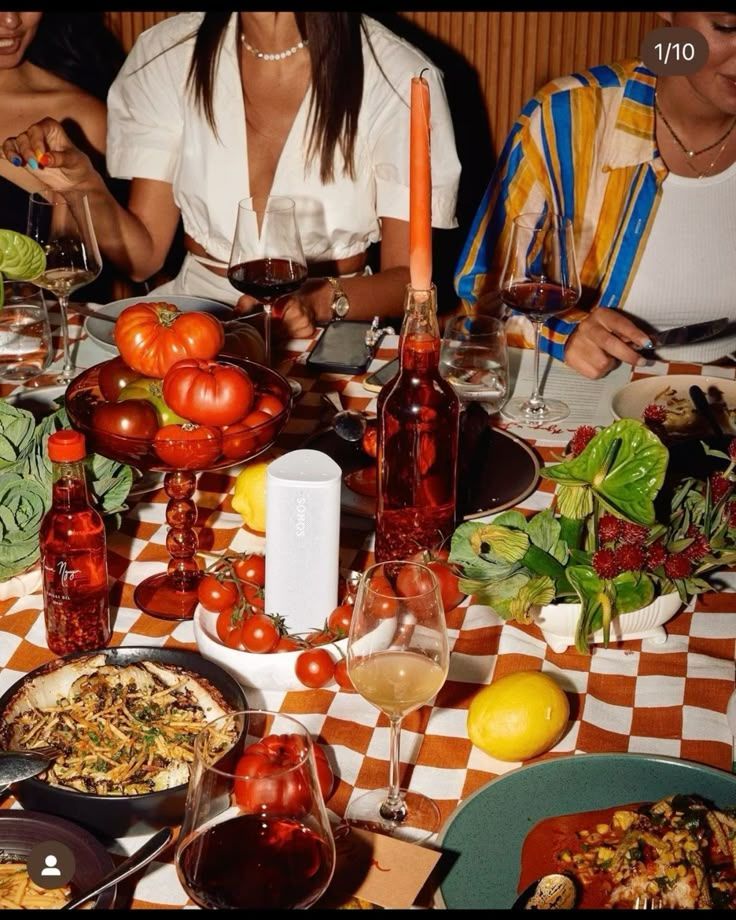 Una mesa de comedor con comida, bebidas y gente. Mantel a cuadros, velas, tomates y verduras.