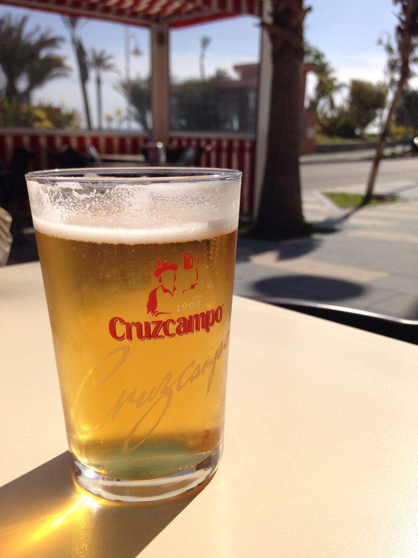 Vaso de cerveza Cruzcampo en una mesa soleada al aire libre. Palmeras y carretera al fondo.